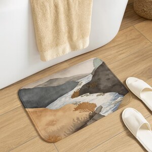 Op de afbeelding: Een rechthoekige badmat met een aquarel landschap van een rivier die door een kloof stroomt. De mat heeft een kleurenpalet van beige, bruin en grijs. Witte slippers liggen naast de mat en een beige handdoek hangt in de buurt.