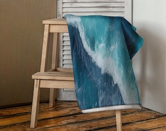 May include: A blue and white ocean wave design throw blanket draped over a wooden chair. The blanket features shades of blue and white, depicting a crashing wave. The chair is made of light-colored wood, and the background includes a wooden floor and a white shutter.