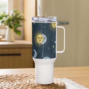 Könnte beinhalten: Ein weiß-blauer Reisebecher mit Henkel und transparentem Deckel. Der Becher hat ein Himmelsdesign mit Sonnen, Monden und Sternen. Ein klarer Strohhalm ist in den Deckel gesteckt. Der Becher steht auf einer gewebten Matte.