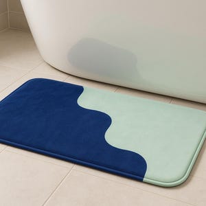以下が含まれることがあります： ツートンカラーのデザインの長方形のバスマット。マットは、濃い青色の部分と明るい緑色の部分の2つのセクションに分かれています。マットの端は丸みを帯びています。マットはタイルの床に置かれています。