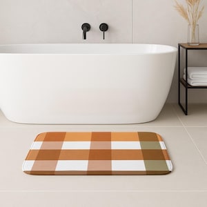 Puede incluir: Una alfombra de baño rectangular con un diseño a cuadros marrón, naranja, blanco y verde. La alfombra está colocada frente a una bañera blanca en un baño moderno. El diseño a cuadros añade un toque de calidez y estilo a la decoración del baño.