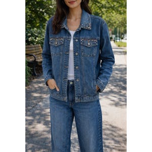 May include: A medium-wash denim jacket and matching jeans. The jacket features embellishments on the collar and pockets. The outfit includes a white top underneath the jacket. The model is standing outside on a sunny day.