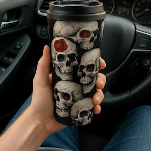May include: Black travel mug featuring a design of multiple skulls with red and white accents. The skulls are arranged in a vertical pattern. The mug has a black lid and is held in a car.