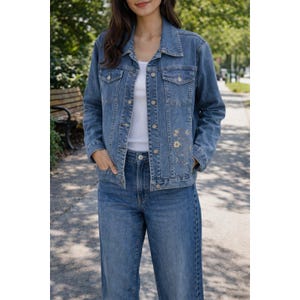 May include: A light blue denim jacket with gold embroidered floral and celestial designs. The jacket has a classic collar, button closures, and front pockets. The model is wearing the jacket with matching blue jeans.