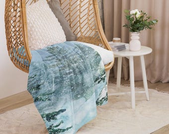 May include: A light blue throw blanket with a winter forest scene. The blanket features a watercolour design of snow-covered trees and a snowy landscape. The blanket is draped over a wicker chair with white and grey pillows.