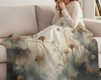 May include: A soft, floral-patterned blanket in shades of beige, brown, and blue. The blanket features a watercolor design of tall, slender stems with flower-like shapes. The blanket is draped over a sofa.