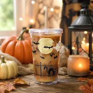Puede incluir: Un vaso de plástico transparente con tapa y pajita, lleno de una bebida helada. El exterior presenta un gráfico con temática de Halloween con una luna llena amarilla, murciélagos negros, un fantasma blanco y un gato negro sobre una valla.