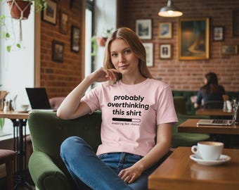 Op de afbeelding: Een lichtroze t-shirt met de tekst "probably overthinking this shirt" in het zwart. Daaronder staat een laadbalkgrafiek en de woorden "analyzing font choice...". Het t-shirt wordt door een persoon gedragen.