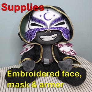May include: A plush doll with an embroidered face, mask, and armor. The doll is wearing a black hooded cloak with gold trim. The mask is white with a purple embroidered design and a crescent moon and stars.