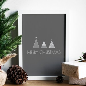 May include: A white framed print with a gray background featuring three stylized Christmas trees and the text "Merry Christmas". The print is sitting on a wooden surface with pine cones and a wrapped gift.