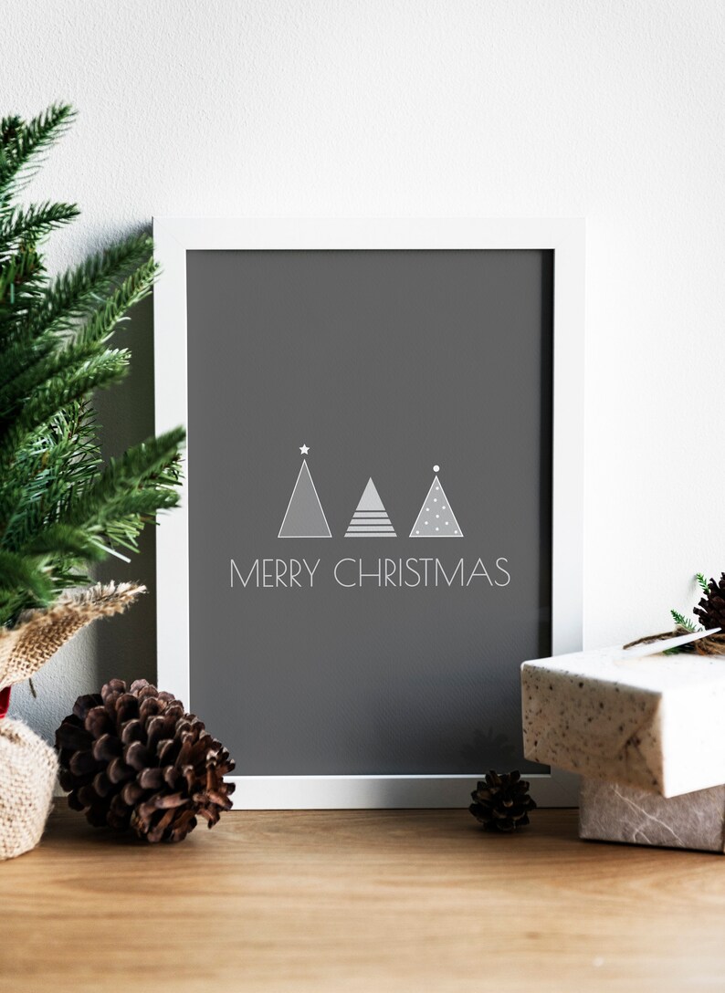 May include: A white framed print with a gray background featuring three stylized Christmas trees and the text "Merry Christmas". The print is sitting on a wooden surface with pine cones and a wrapped gift.