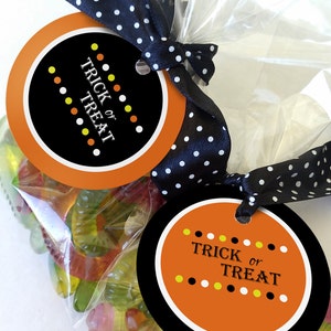 May include: Two round orange and black Halloween treat bags with a black and white polka dot ribbon. The bags are filled with gummy worms and have tags that say "Trick or Treat" with yellow and white polka dots.