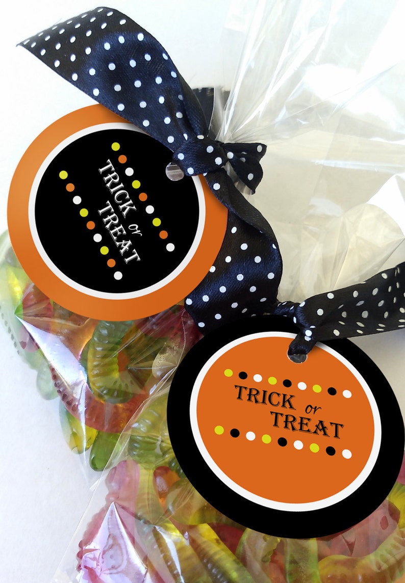 May include: Two round orange and black Halloween treat bags with a black and white polka dot ribbon. The bags are filled with gummy worms and have tags that say "Trick or Treat" with yellow and white polka dots.