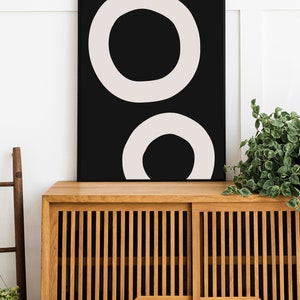 Puede incluir: Una impresi&oacute;n de arte abstracto en blanco y negro con dos c&iacute;rculos conc&eacute;ntricos. La impresi&oacute;n est&aacute; enmarcada y colgada en una pared blanca sobre un armario de madera.