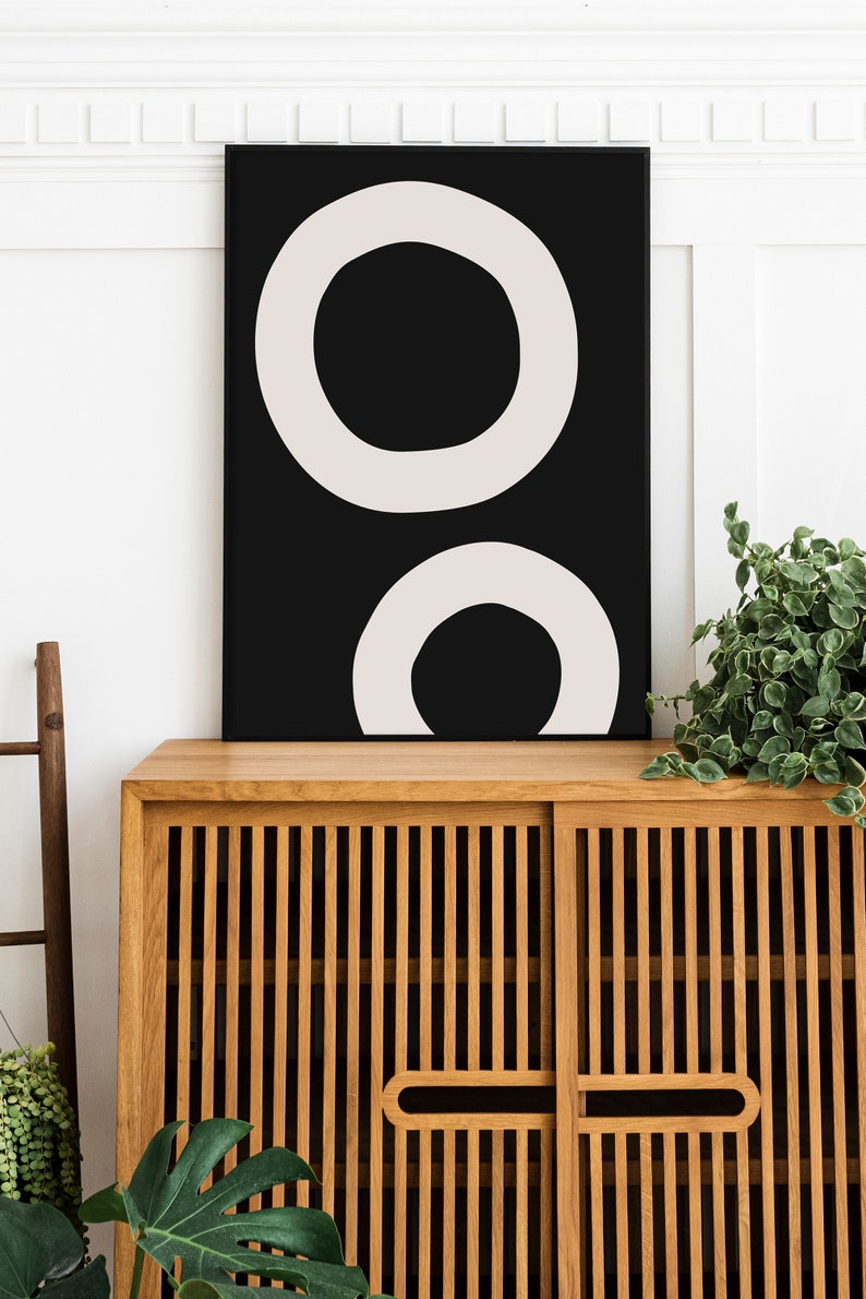 Puede incluir: Una impresi&oacute;n de arte abstracto en blanco y negro con dos c&iacute;rculos conc&eacute;ntricos. La impresi&oacute;n est&aacute; enmarcada y colgada en una pared blanca sobre un armario de madera.