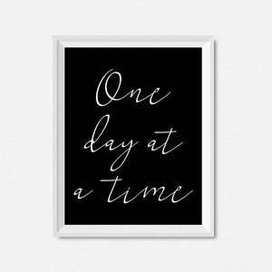May include: Black and white framed print with the text "One day at a time" in white cursive script.