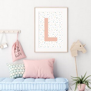May include: A framed print featuring a pink letter L on a white background with colourful polka dots. The print is hanging on a white wall above a light blue tufted cushion.