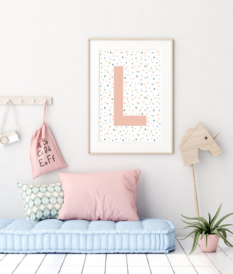 May include: A framed print featuring a pink letter L on a white background with colourful polka dots. The print is hanging on a white wall above a light blue tufted cushion.