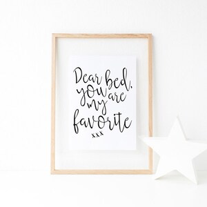 May include: A framed print with the text "Dear bed, you are my favorite xxx" in black cursive font on a white background. The frame is light wood and there is a white star on the table below the frame.