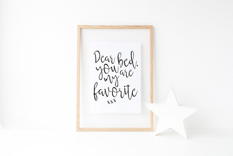 May include: A framed print with the text "Dear bed, you are my favorite xxx" in black cursive font on a white background. The frame is light wood and there is a white star on the table below the frame.