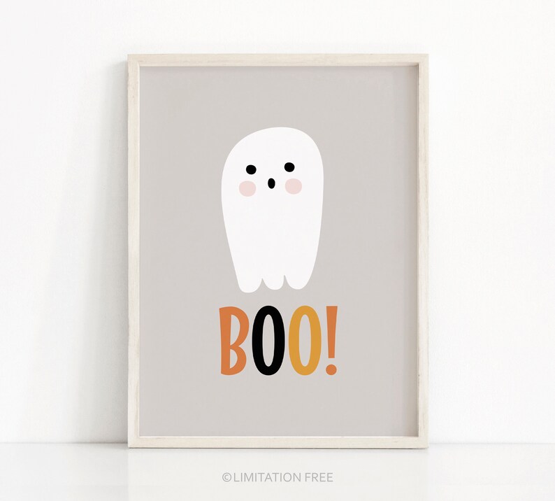 May include: A framed print featuring a white cartoon ghost with a blush pink face and black eyes. Below the ghost, the word "BOO!" is written in orange, black, and yellow. The background is a light gray color.