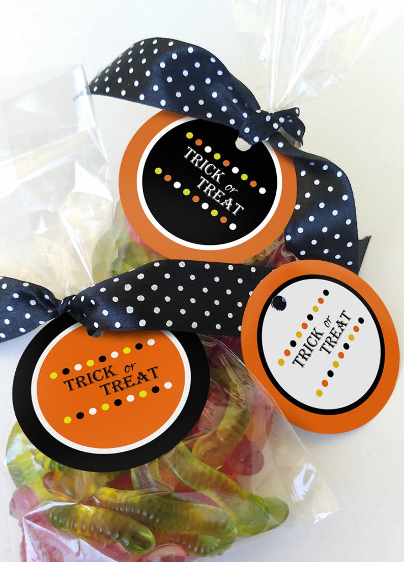May include: Two round orange and black Halloween treat bags with white polka dot ribbon. The bags are filled with gummy worms and have tags that say "Trick or Treat" with orange, yellow, and white polka dots.