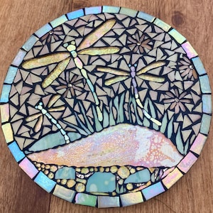 May include: A round mosaic wall art featuring a design of four iridescent dragonflies with a background of small, irregular, black and white tiles. The dragonflies are made of iridescent glass tiles and are arranged in a circular pattern. The background tiles are arranged in a random pattern. The mosaic is set on a light brown wooden surface.