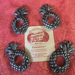 May include: Four silver pineapple napkin rings. The rings are made of pewter and have a detailed design. The rings are on a red tablecloth with a card that reads "Olde Country Reproductions Built 1823 Pewtarex™ Hand Finished Metalware from York, Pennsylvania 722 West Market Street P.O. Box 2617 York, PA 17405 (717) 848-1859"