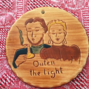 May include: A round, wooden plaque with a painted scene of two people holding a candle. The plaque has the words "Quen the light" written on it.