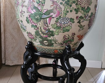 May include: A large, round, white porcelain planter with a black wooden stand. The planter is decorated with a floral pattern featuring peacocks, flowers, and leaves in shades of green, pink, and yellow.