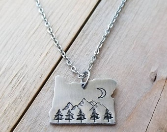 May include: A silver-colored pendant necklace featuring the outline of the state of Oregon. The pendant has a mountain and tree design with a crescent moon. The necklace is on a silver chain.
