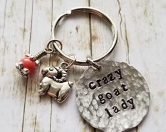 May include: A silver-toned keychain with a round tag that reads "Crazy goat lady." The keychain also features a goat charm and a red bead charm. The tag has a hammered texture.