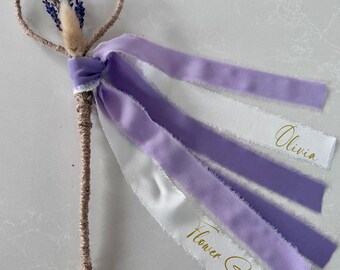 May include: A heart-shaped wand with a woven handle and dried lavender and bunny tail accents. The wand has flowing white and lilac ribbons, with the words "Olivia" and "Flower Girl" printed in gold.