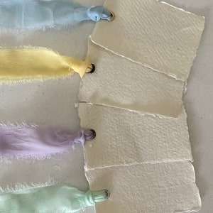 Tarjetas de lugar de boda de lujo con cinta pastel, arreglos de mesa en blanco con bordes sin rematar y tarjetas de nombre con cinta de gasa deshilachada a mano para la mesa de boda.