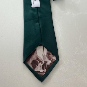 May include: A dark green tie with a satin finish. The tie has a white label with the word "olive" printed on it. The tip of the tie features a black and white photograph of a couple.