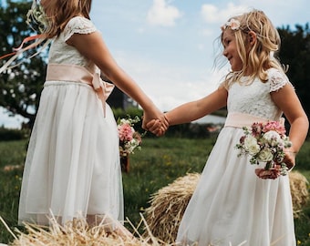 May include: Two young girls in white dresses with pink sashes and floral headbands hold hands and smile while standing in a field of hay bales.
