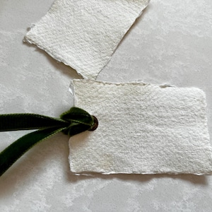 May include: A white, textured paper tag with a green velvet ribbon tied through a brass eyelet. The tag has a slightly irregular shape.