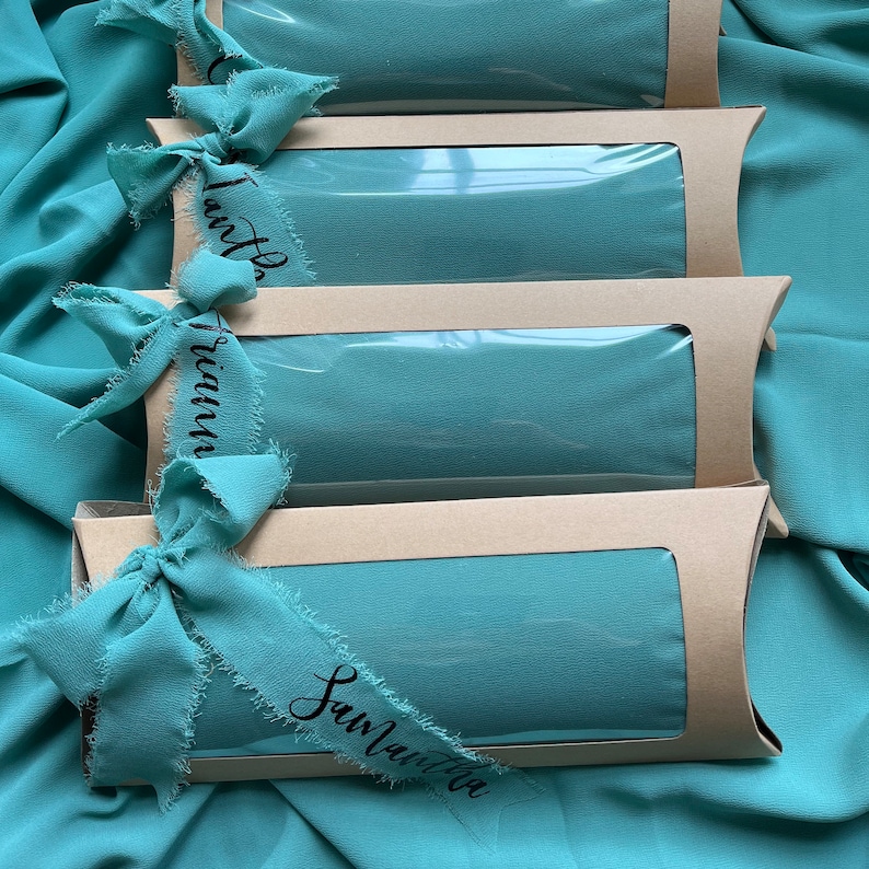 May include: Four teal blue scarves are packaged in brown boxes with clear plastic windows. Each box is tied with a teal blue ribbon with a frayed edge and a handwritten tag. The tags read "Lautha", "Triann", and "Sawlaitta".