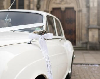 May include: A cream-coloured vintage car, adorned with a white ribbon and bow, is parked near a church. A white ribbon on the side mirror reads "Just Married 2.08.2026". The car has a classic design with chrome details, suitable for a wedding.