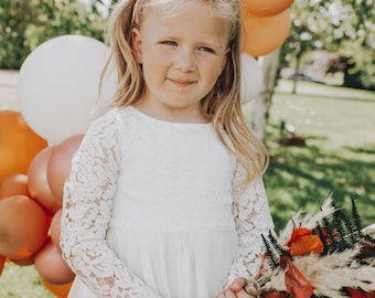Op de afbeelding: Een jong meisje in een witte kanten jurk houdt een boeket gedroogde bloemen vast met bruine, oranje en groene accenten. Ze staat voor een achtergrond van oranje en witte ballonnen.