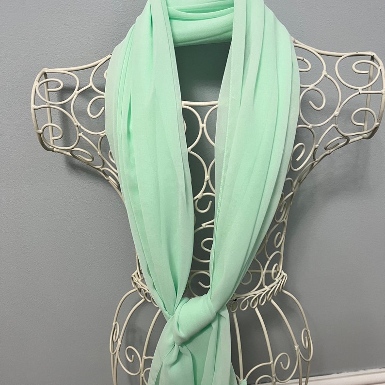 May include: A light green chiffon scarf draped over a white wire mannequin. The scarf is folded in half and tied in a knot at the front.