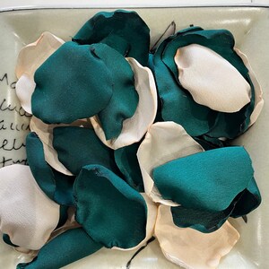 Op de afbeelding: Een verzameling teal en ivoor zijden rozenblaadjes. De blaadjes zijn willekeurig gerangschikt op een lichtbruin oppervlak.
