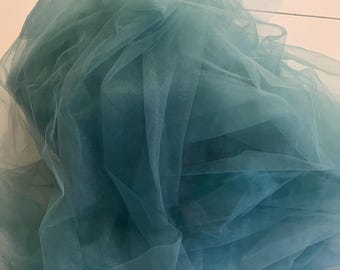 May include: A pile of teal tulle fabric. The fabric is layered and appears soft and delicate. The colour varies slightly throughout the fabric, creating depth and visual interest. The tulle is likely used for crafting or decorative purposes.