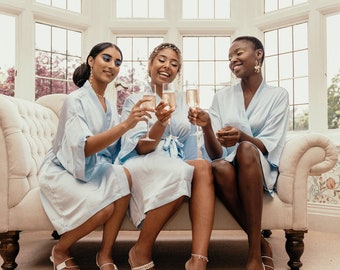 May include: Three people wearing light blue satin robes are sitting on a white couch and holding champagne flutes. They are smiling and looking at each other.