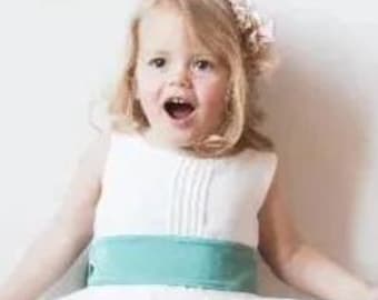 May include: A young child in a white dress with a teal sash. The dress features a pleated bodice and a full skirt. The child has blonde hair and wears a floral hair accessory. The child is smiling with their mouth open.