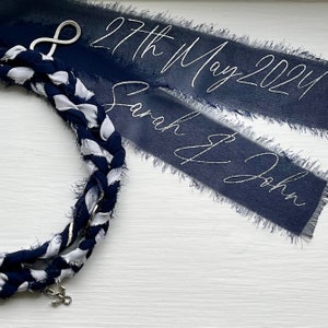 May include: A navy blue and white braided rope with an infinity symbol charm and an anchor charm. Two navy blue ribbon banners with white text read "27th May 2021" and "Sarah & John".