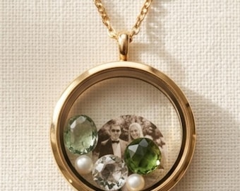 May include: A gold-tone circular floating locket pendant on a matching chain. The locket contains a black and white photograph of two people, two faceted green gemstones, one faceted clear gemstone, and two small white spherical pearls inside.