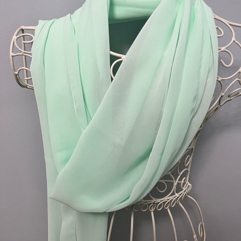 May include: A light green chiffon scarf draped over a white metal display stand. The scarf is soft and flowing, with a delicate texture.