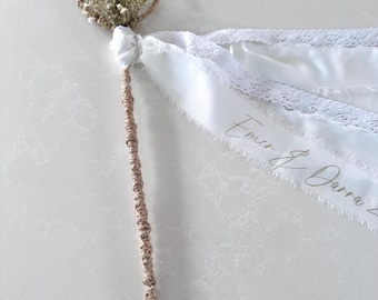 May include: A wedding wand featuring a heart-shaped top made of woven material, decorated with small dried flowers. The handle is wrapped in a similar woven material. White ribbons with lace trim and the text "Emer & Darra" are attached.