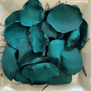 Op de afbeelding: Een verzameling tealgroene zijden bloemblaadjes, gerangschikt in een kom. De bloemblaadjes zijn zacht en delicaat, met een lichte glans.